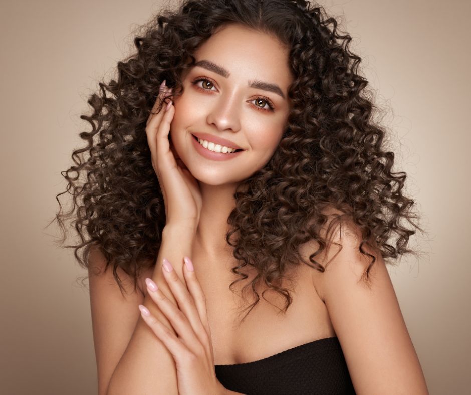 A joyful young woman with lush, curly hair and a bright smile, gently cradling her cheek with her hand.